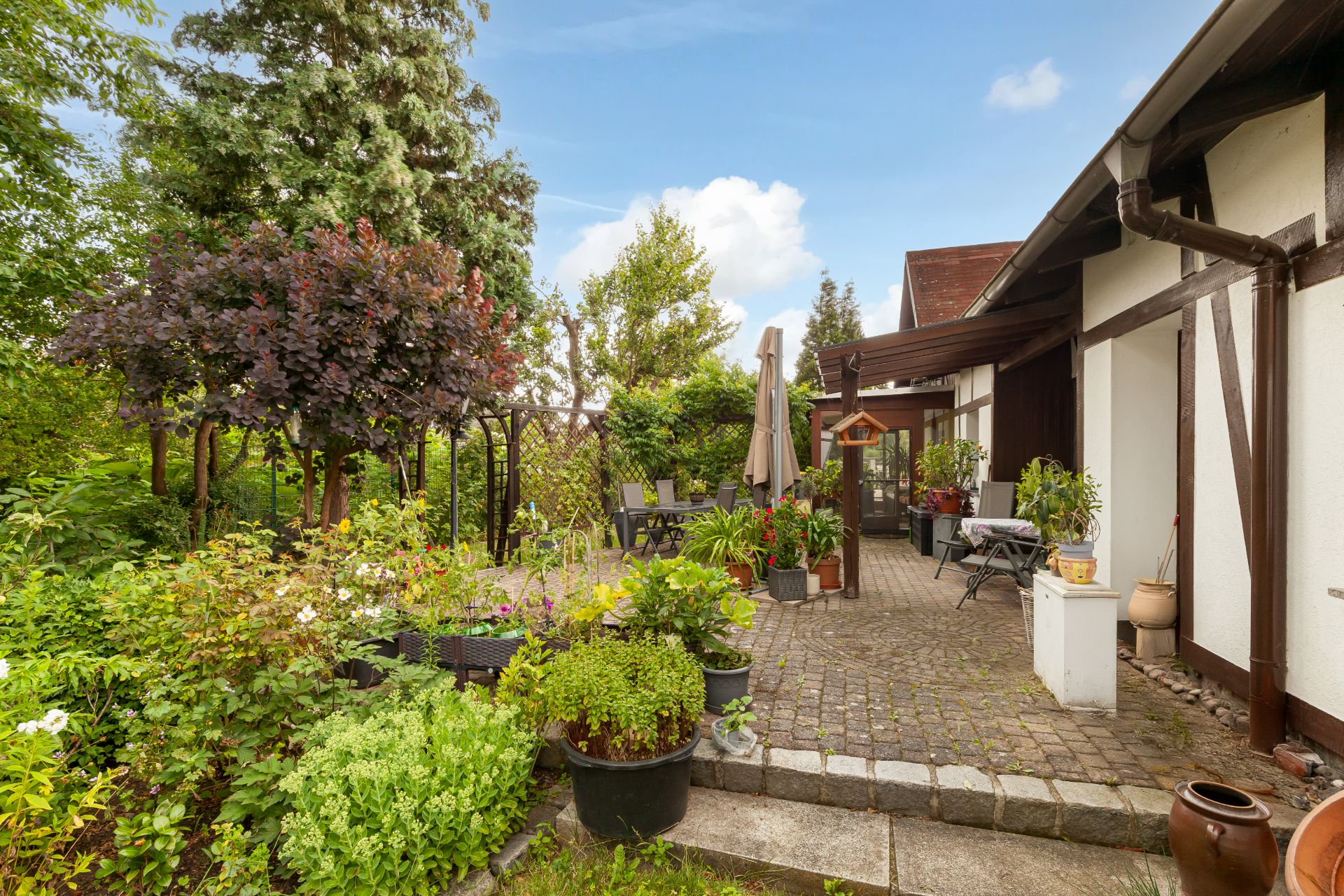 Gepflasterte Terrasse mit Pflanzen, Gartenmöbeln und Sonnenschirm neben einem Wohnhaus.