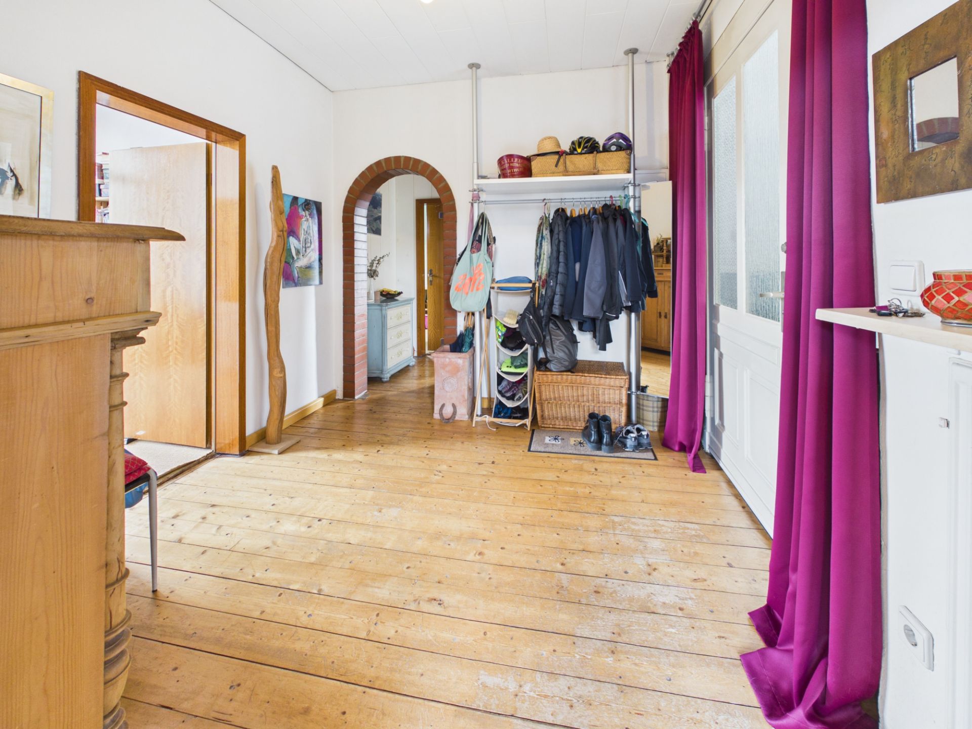 Flur mit Holzboden, Garderobe, Jacken, Schuhen und lila Vorhängen an der Tür.