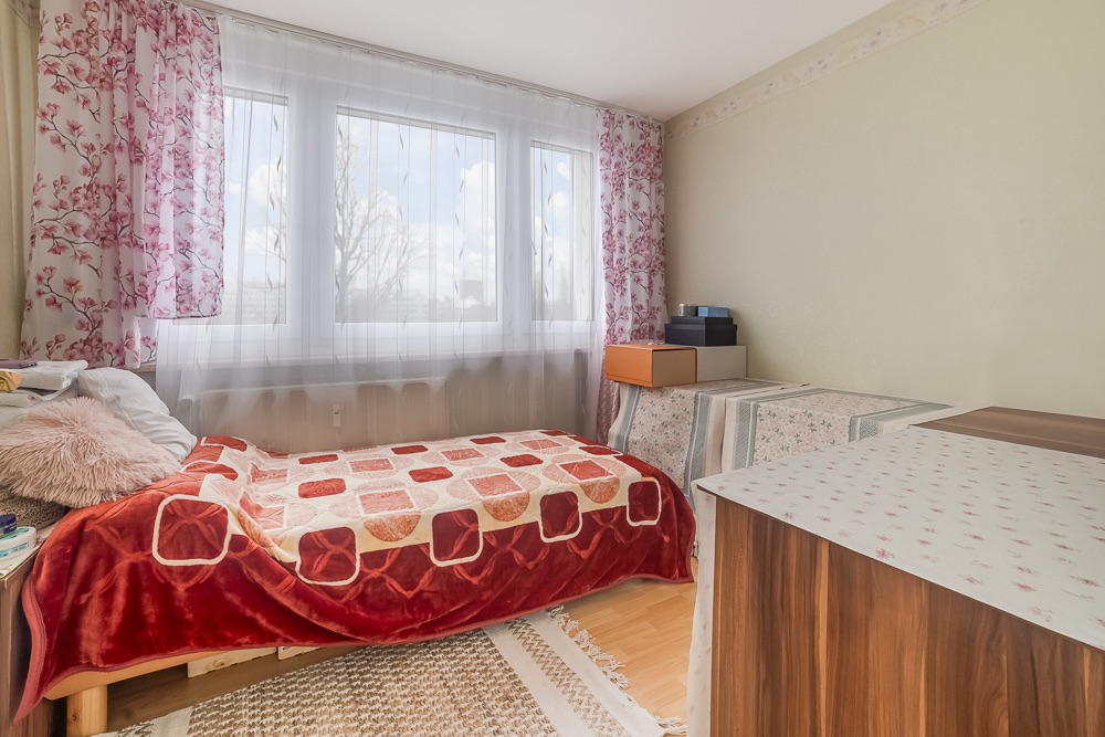 Helles Schlafzimmer mit Einzelbett, bunten Decken, Teppich und großem Fenster mit Blumenvorhängen.