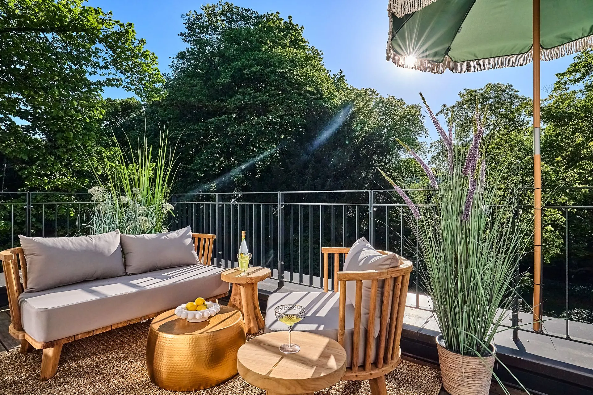 Sonnige Terrasse mit Sitzmöbeln, kleinen Tischen, Pflanzen und Sonnenschirm, umgeben von Bäumen.