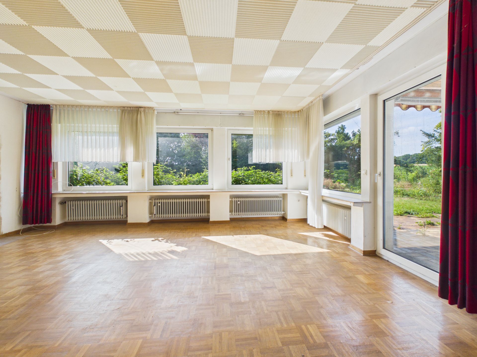 Heller, leerer Raum mit Parkettboden, großen Fenstern und Vorhängen, Blick auf Garten.