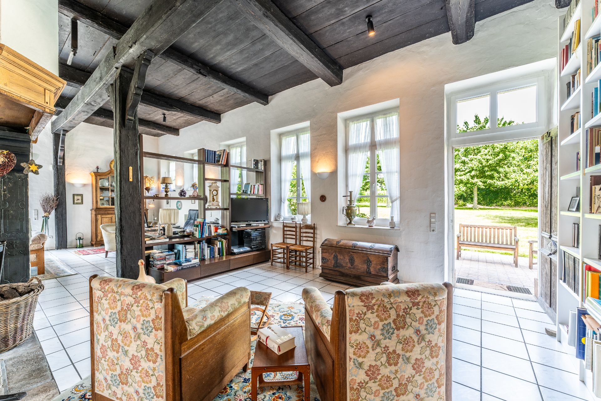 Helles Wohnzimmer mit Holzbalken, zwei Sesseln, Bücherregal und Zugang zu einem Garten.
