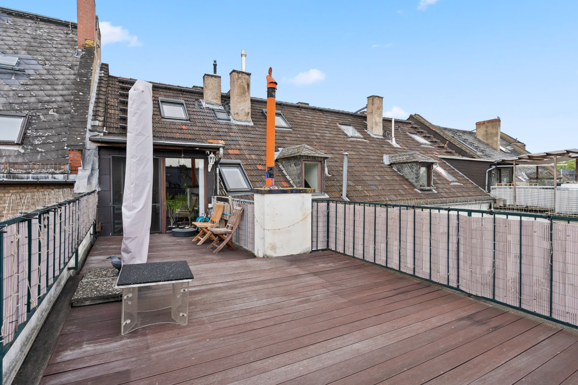 Dachterrasse mit Holzboden, Tisch, Stühlen und Sonnenschirmen, umgeben von einem Zaun.