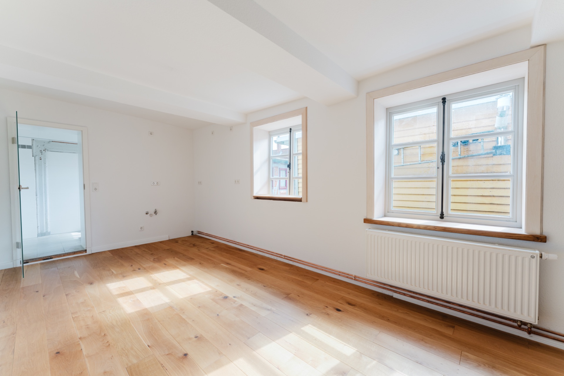 Helles, leeres Zimmer mit Holzboden, zwei Fenstern und einem Heizkörper unter einem Fenster.