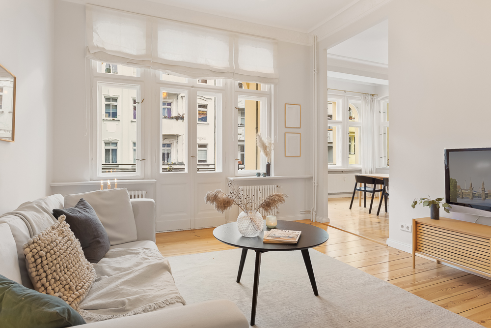 Helles Wohnzimmer mit Sofa, Couchtisch, TV und großen Fenstern, Holzboden und Blick in Esszimmer.