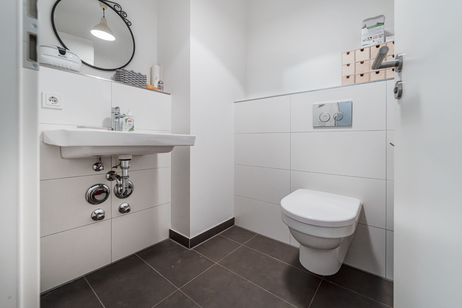 Modernes Bad mit weißem Waschbecken, Spiegel und Wandtoilette auf grauen Fliesen.