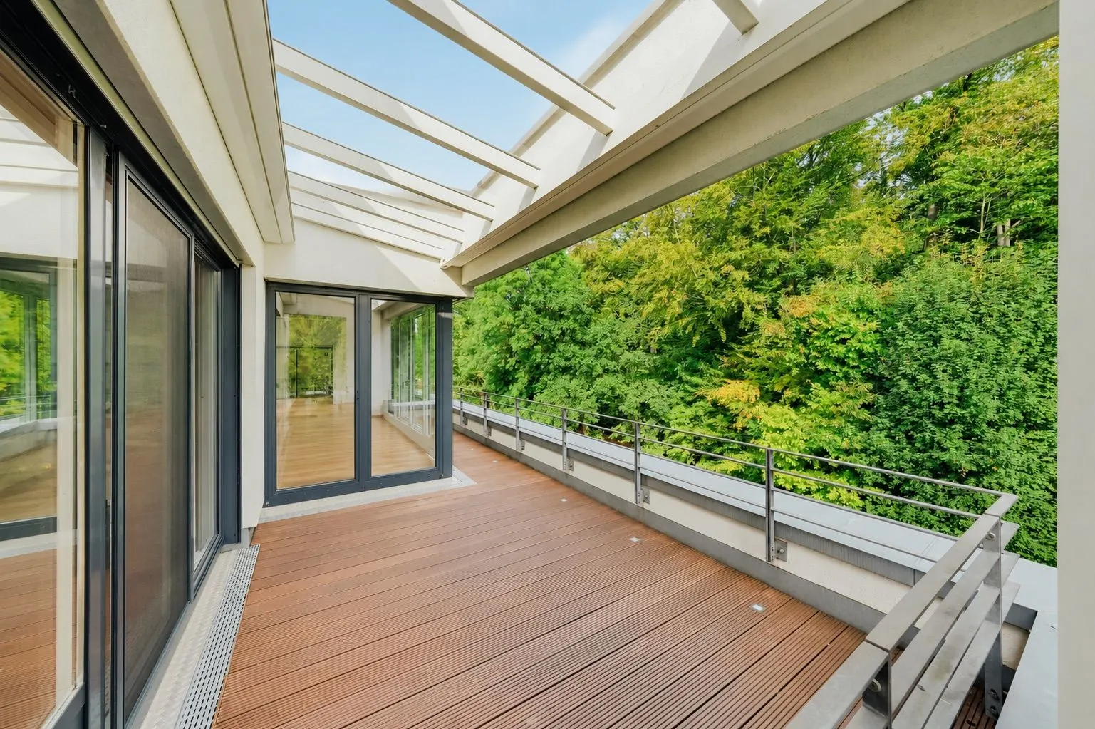 Große Terrasse mit Holzboden, Glasgeländer und Blick auf einen grünen Wald.