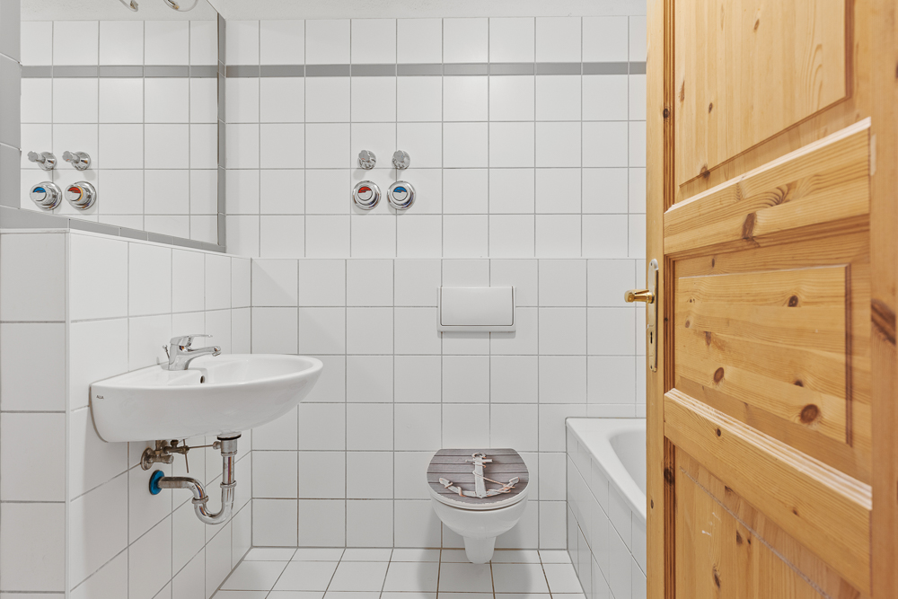 Kleines Badezimmer mit Waschbecken, Toilette und Badewanne, weiße Wandfliesen, Holztür offen.