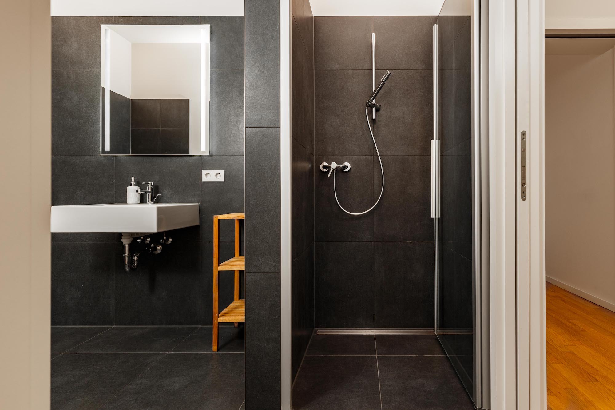 Modernes Bad mit dunklen Fliesen, Dusche, Waschbecken, Spiegel und kleinem Regal.