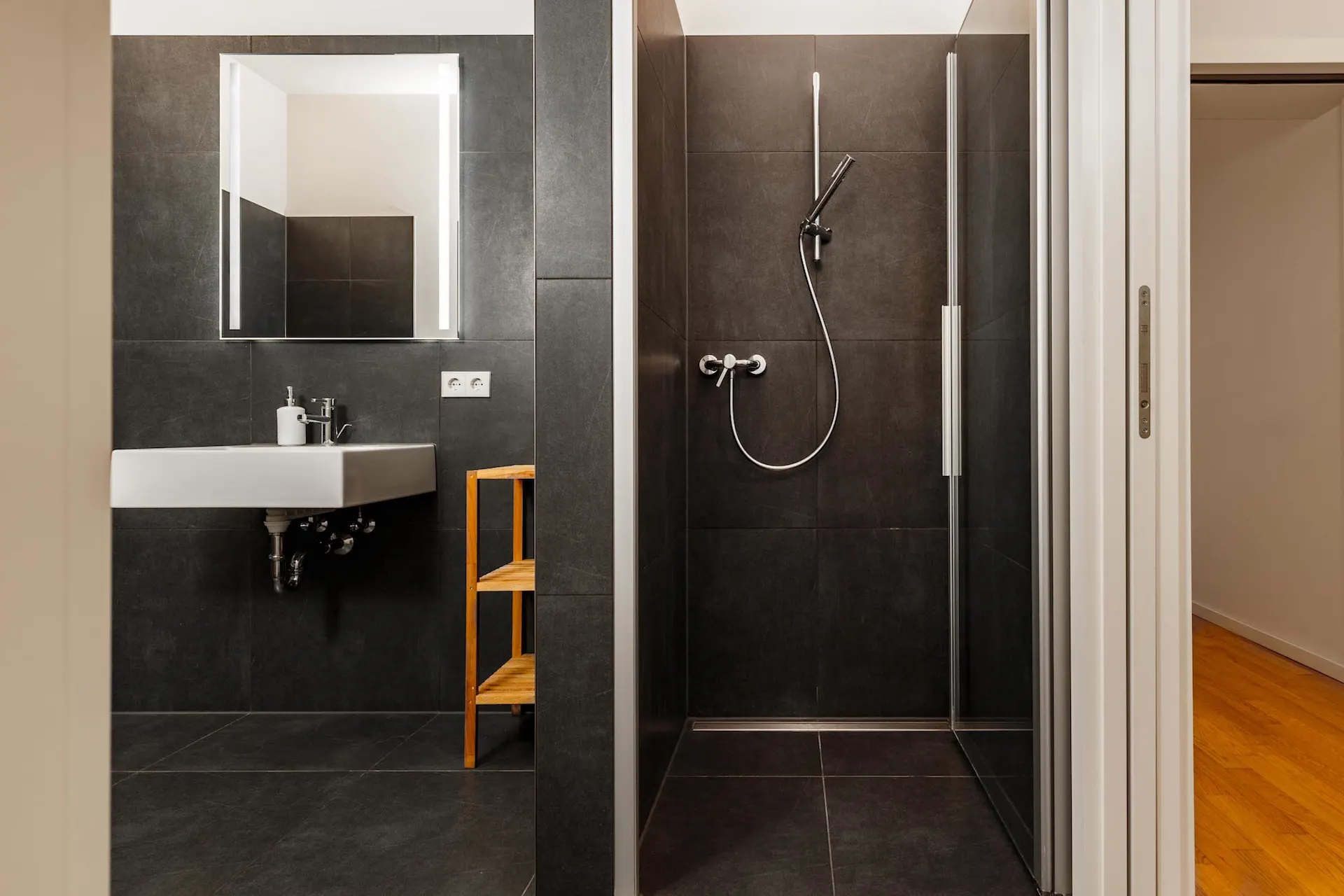 Modernes Bad mit dunklen Fliesen, Dusche, Waschbecken, Spiegel und kleinem Regal.