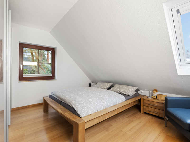 Schlafzimmer mit Holzbett, Nachttisch, Sessel und zwei Fenstern unter einer Dachschräge.