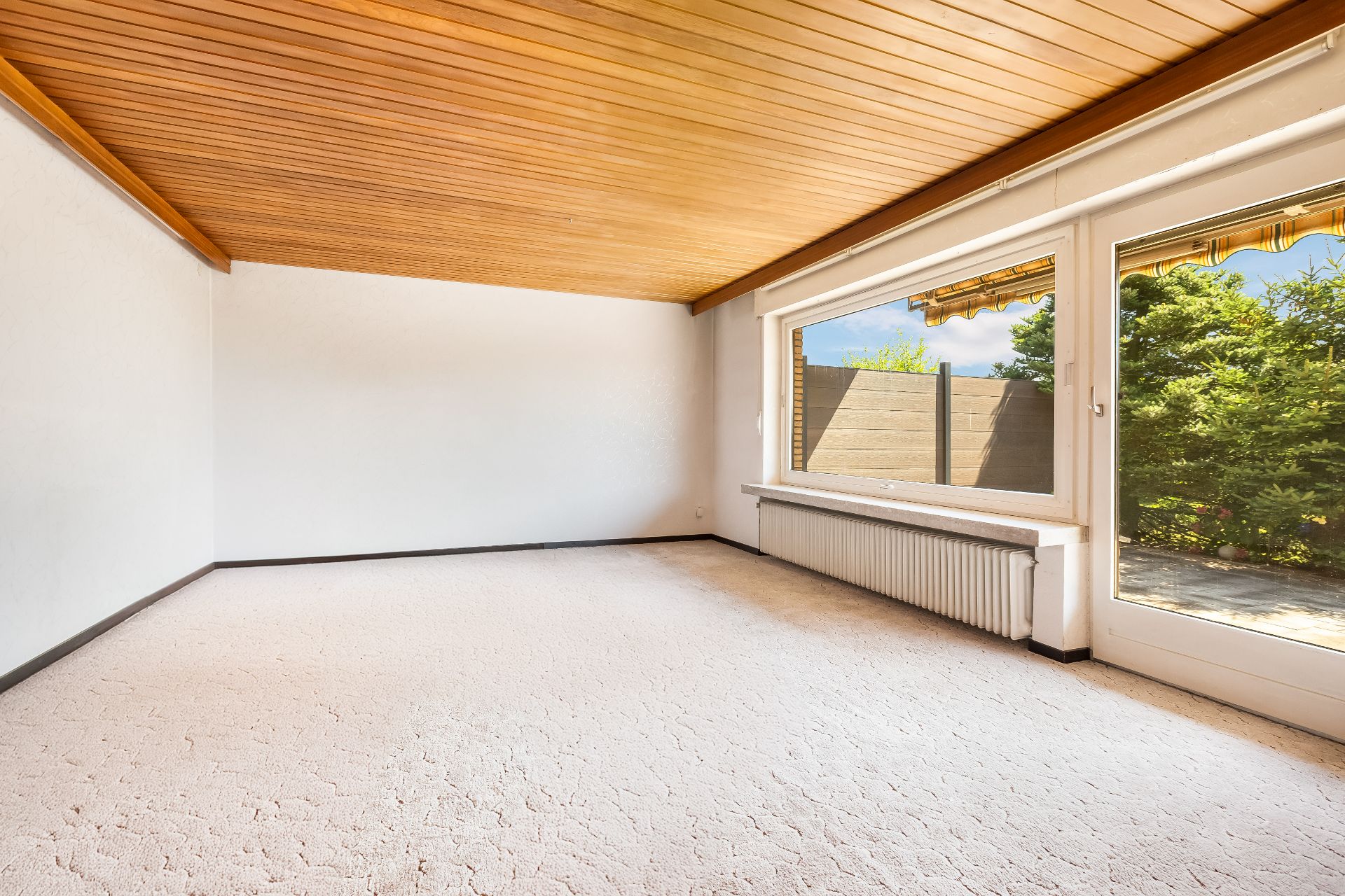 Leerer heller Raum mit Holzdecke, Teppichboden und großem Fenster zur Terrasse.