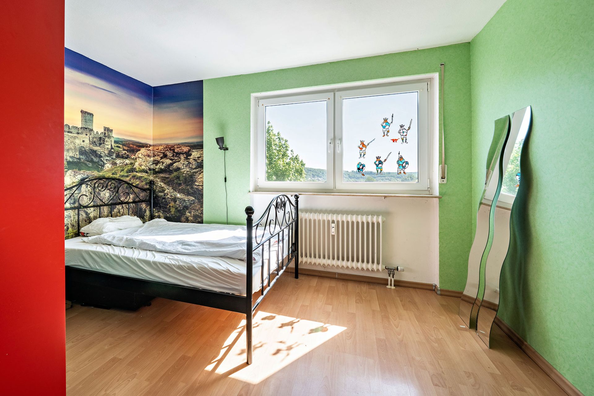 Helles Schlafzimmer mit grünem Anstrich, Einzelbett, Wandbild, großem Fenster und zwei welligen Spiegeln.