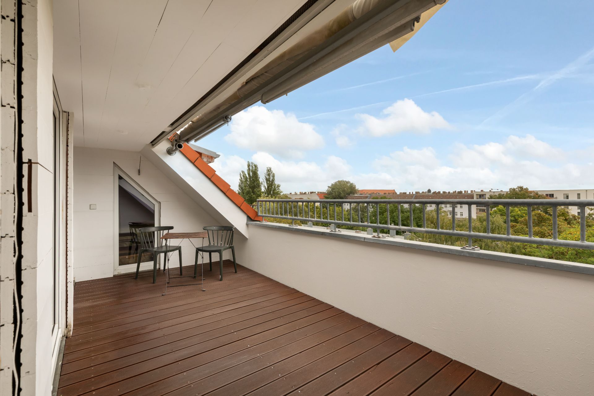 Überdachter Balkon mit Holzboden, zwei Stühlen und Tisch, Blick auf Bäume und Wohngebäude.
