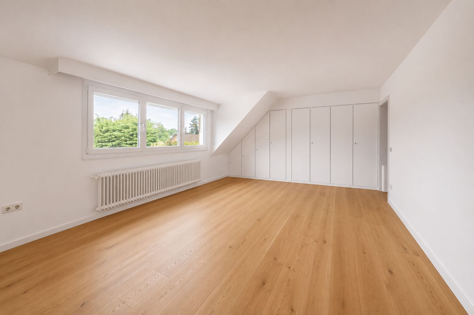 Helles leeres Zimmer mit Holzfußboden, großen Fenstern, Heizkörper und weißen Einbauschränken.
