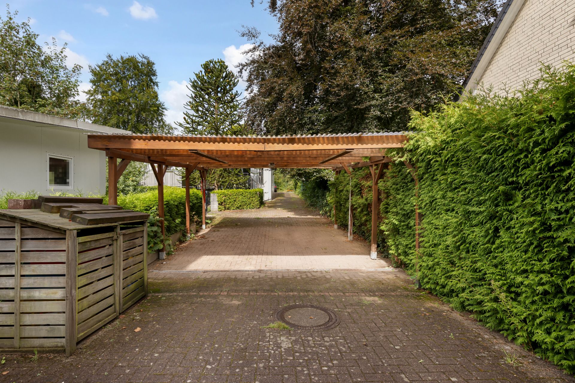 Überdachter Carport mit Holzstruktur neben Hecken und Wohnhäusern, Weg führt hindurch.