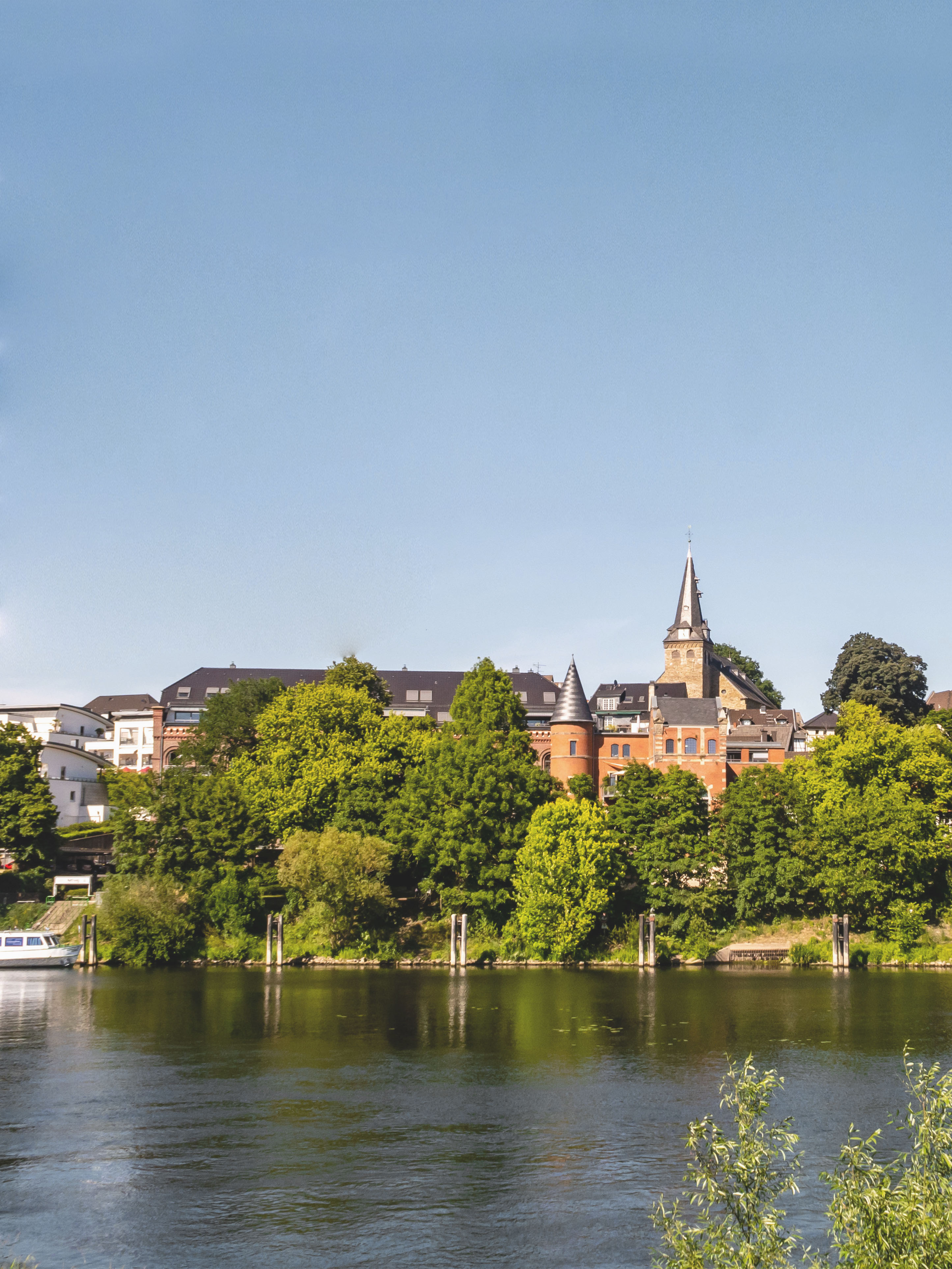 Gebäude und Kirchturm hinter grünen Bäumen an einem Fluss bei klarem Himmel.