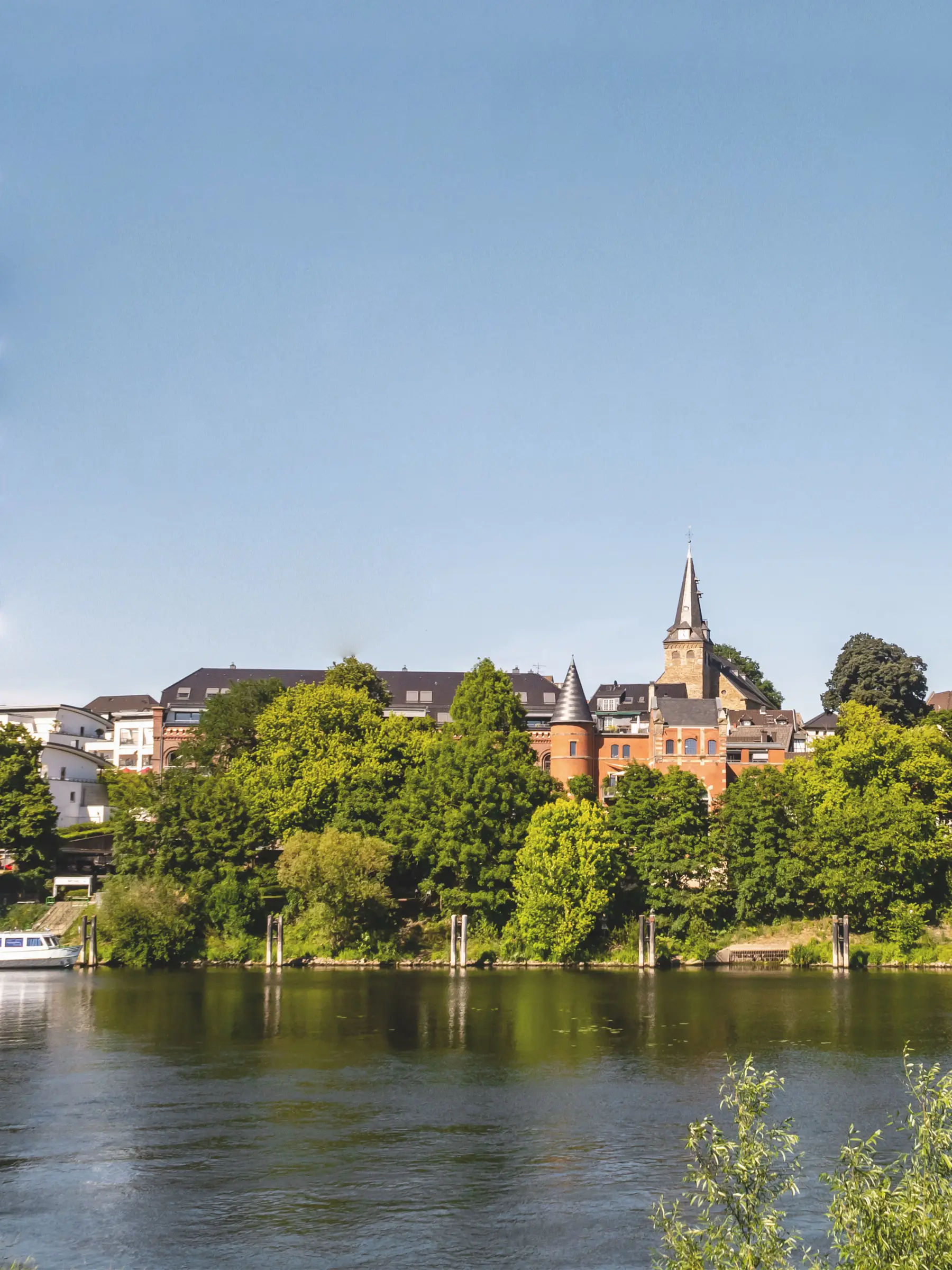 Gebäude und Kirchturm hinter grünen Bäumen an einem Fluss bei klarem Himmel.
