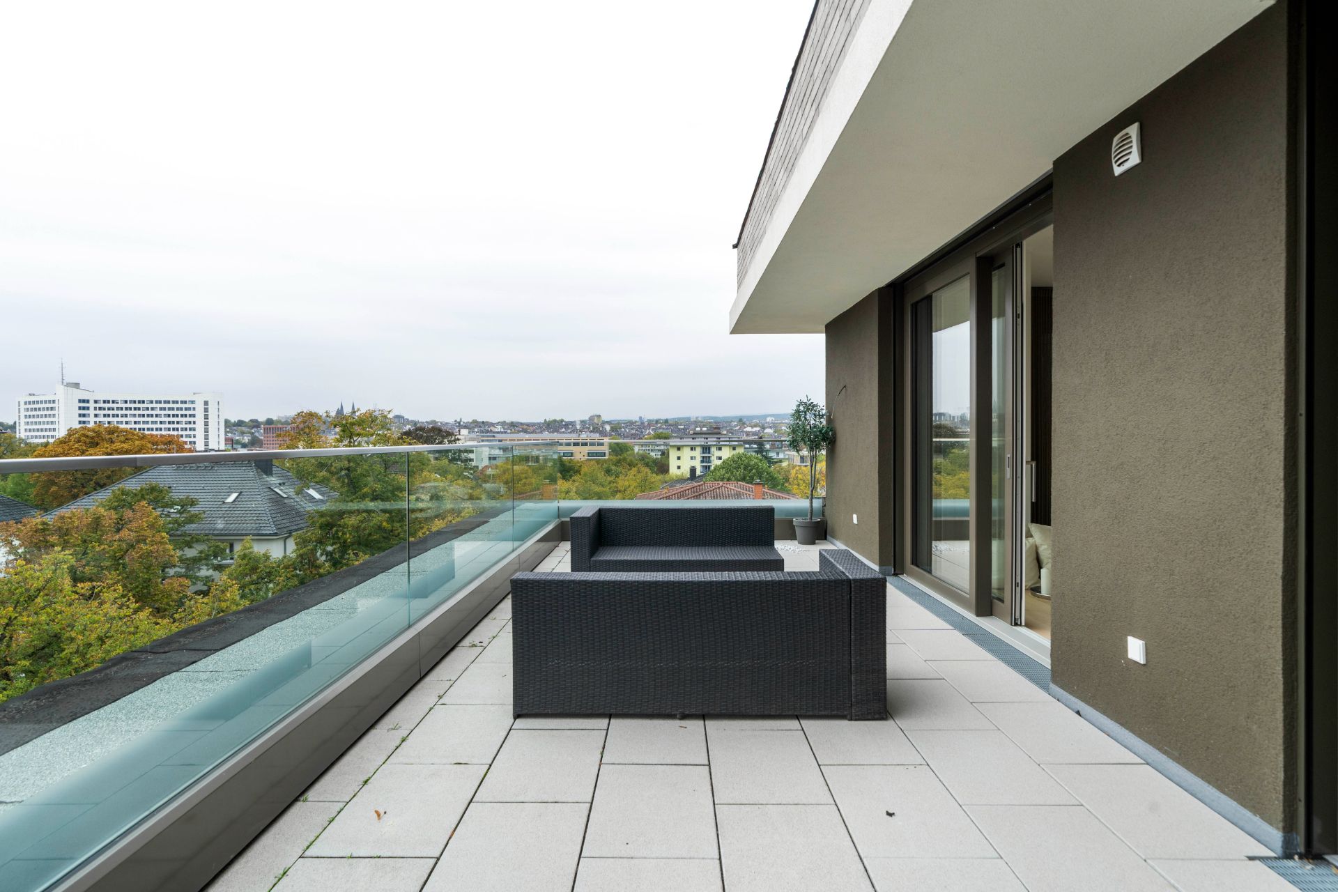 Moderne Terrasse mit Glasgeländer, Sitzgruppe und Blick auf Bäume und Stadt.