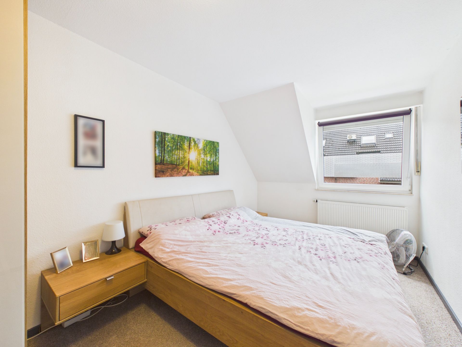 Helles Schlafzimmer mit Doppelbett, Nachttisch, Lampe, Bildern und Fenster mit Blick nach draußen.