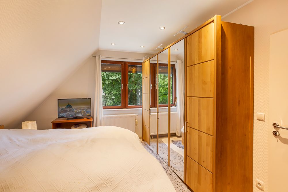 Schlafzimmer mit Holzbett, Kleiderschrank mit Spiegeln und Fenstern mit weißen Vorhängen.