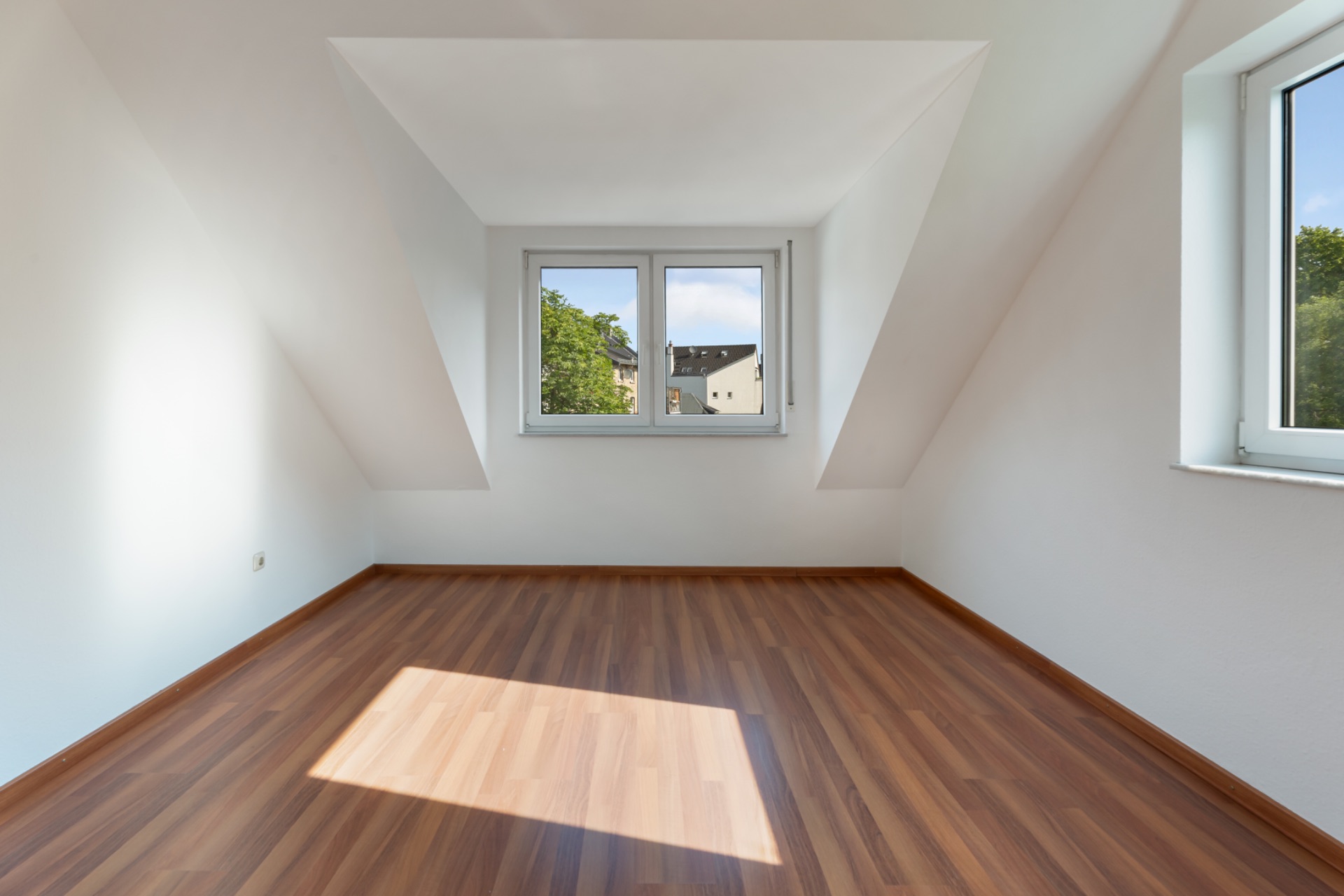 Leerer Dachgeschossraum mit Laminatboden und zwei Fenstern, durch die Tageslicht hereinfällt.