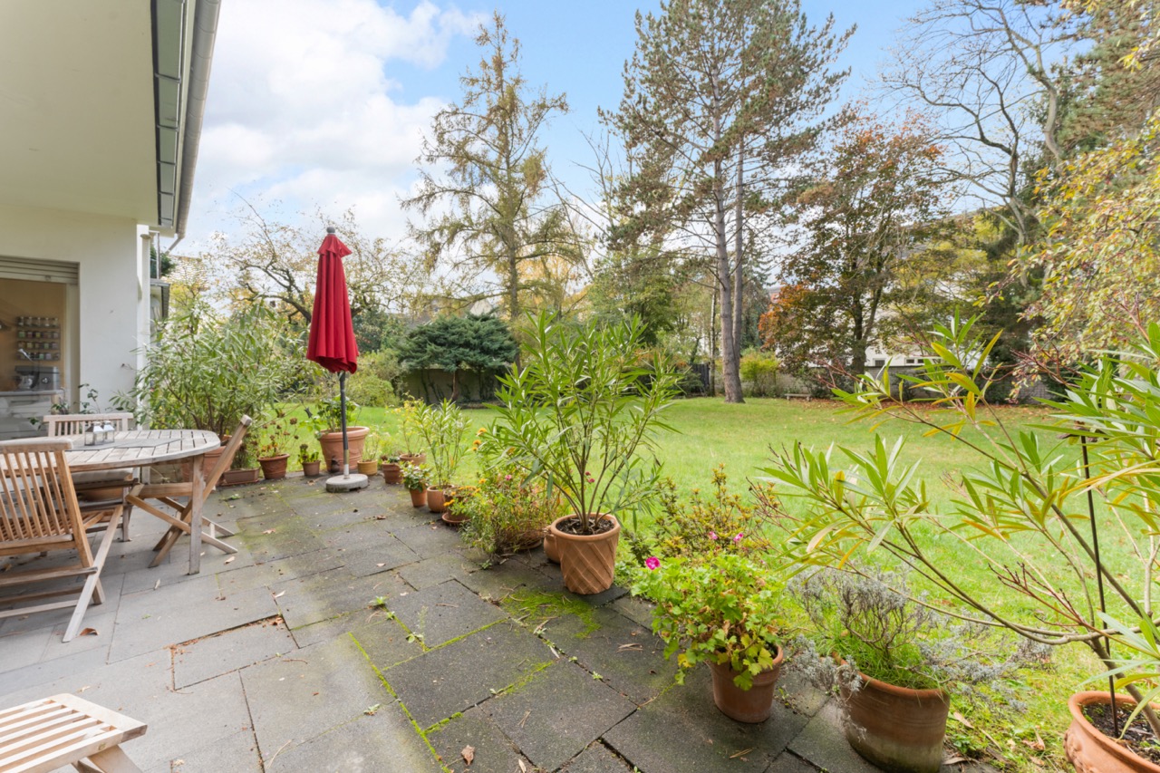 Terrasse mit Holztisch, Stühlen, Blumentöpfen und rotem Sonnenschirm, Blick auf Garten mit Rasen und Bäumen.