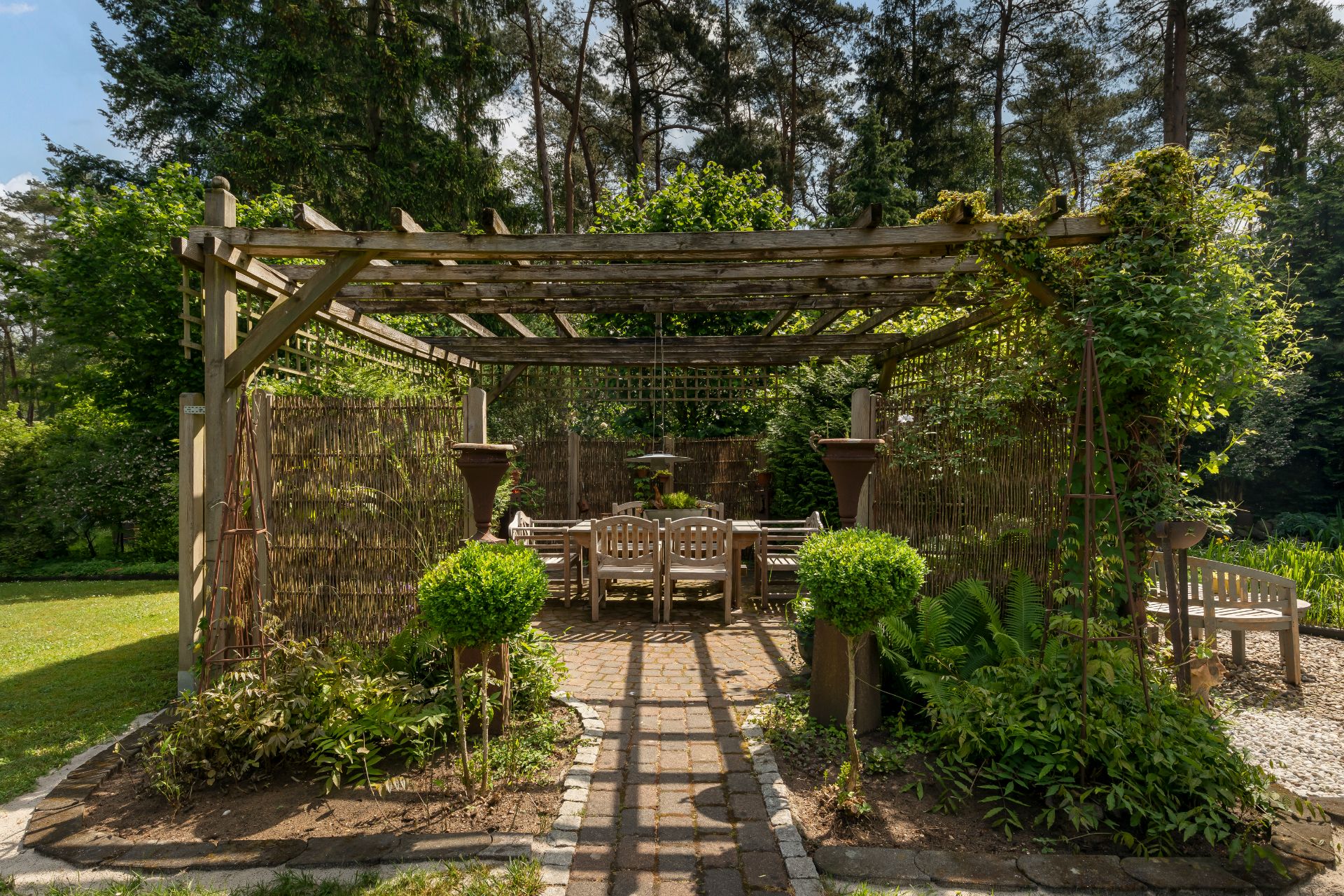 Holzpergola im Garten mit Sitzgruppe und grünen Pflanzen, umgeben von Bäumen.