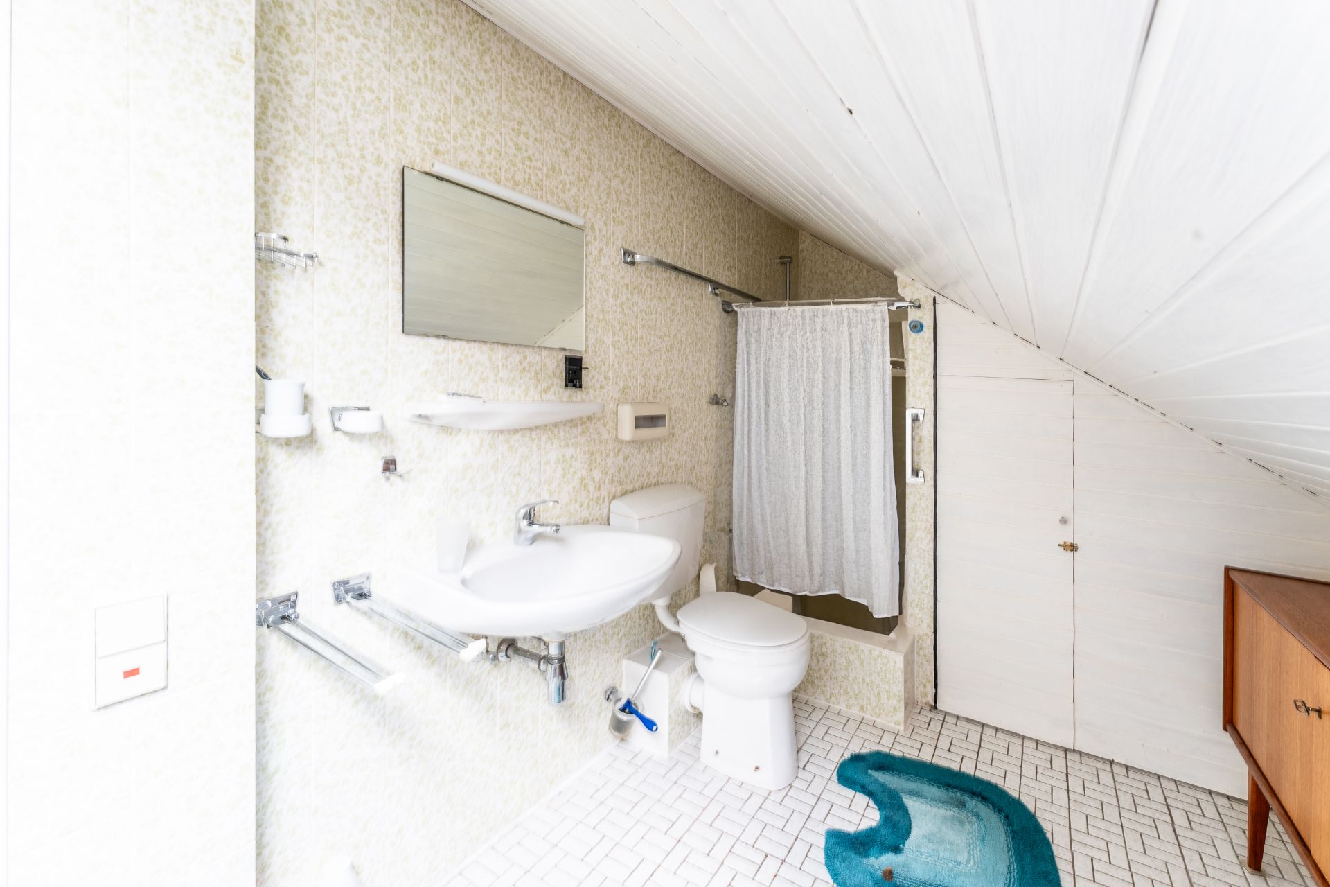 Helles Badezimmer mit Waschbecken, WC, Spiegel, Dusche mit Vorhang und türkisfarbener Badematte.