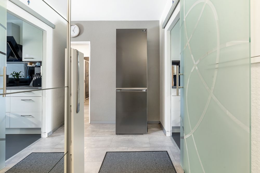 Moderner Flur mit zwei Türen, rechts und links eine Küche, geradeaus ein grauer Kühlschrank.