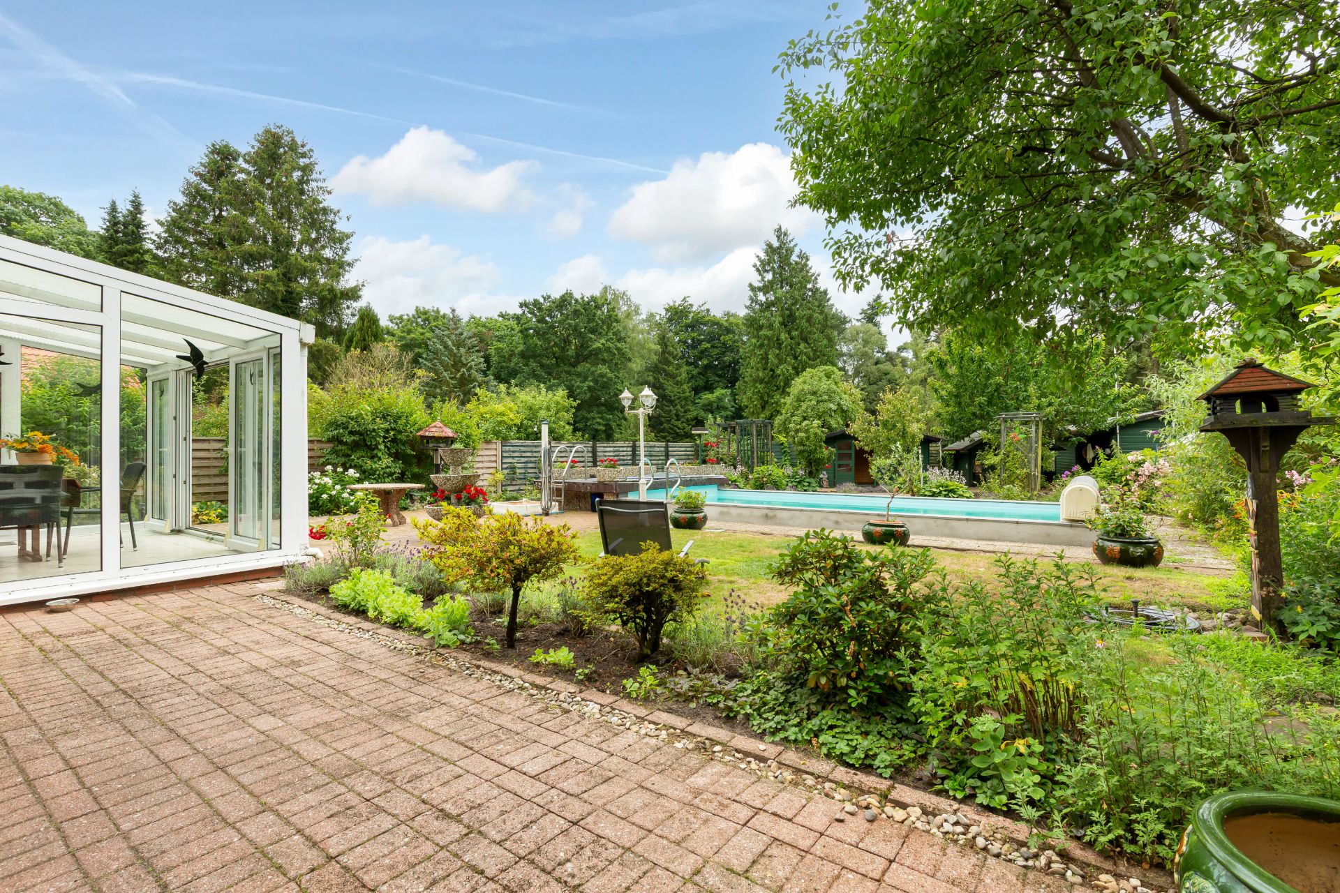 Garten mit Terrasse, Glas-Wintergarten und Swimmingpool, umgeben von Bäumen und Pflanzen.