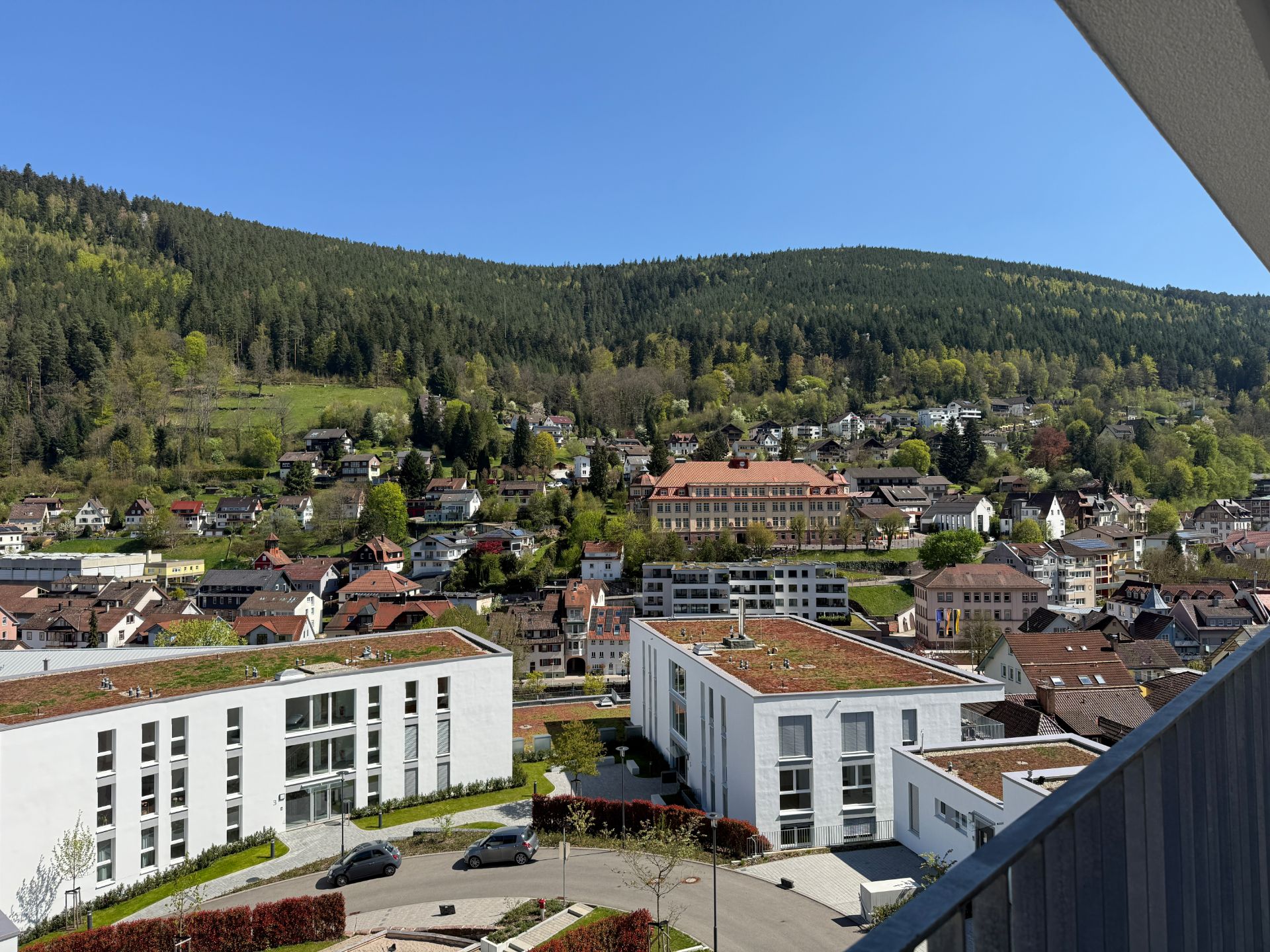 Blick von einem Balkon auf moderne Wohngebäude und eine hügelige, bewaldete Landschaft.