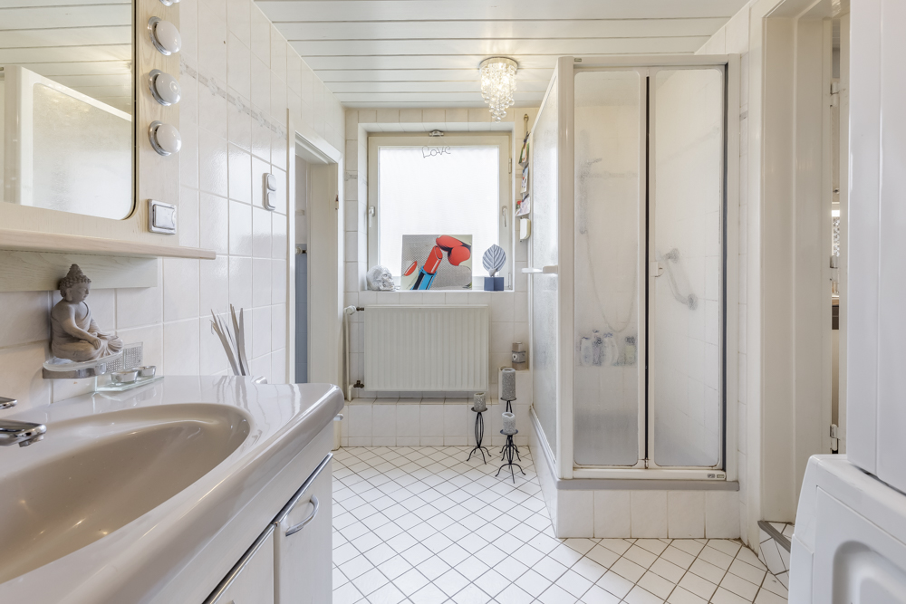 Modernes Badezimmer mit Waschbecken, Dusche, Fenster und Dekorationen auf weißem Fliesenboden.