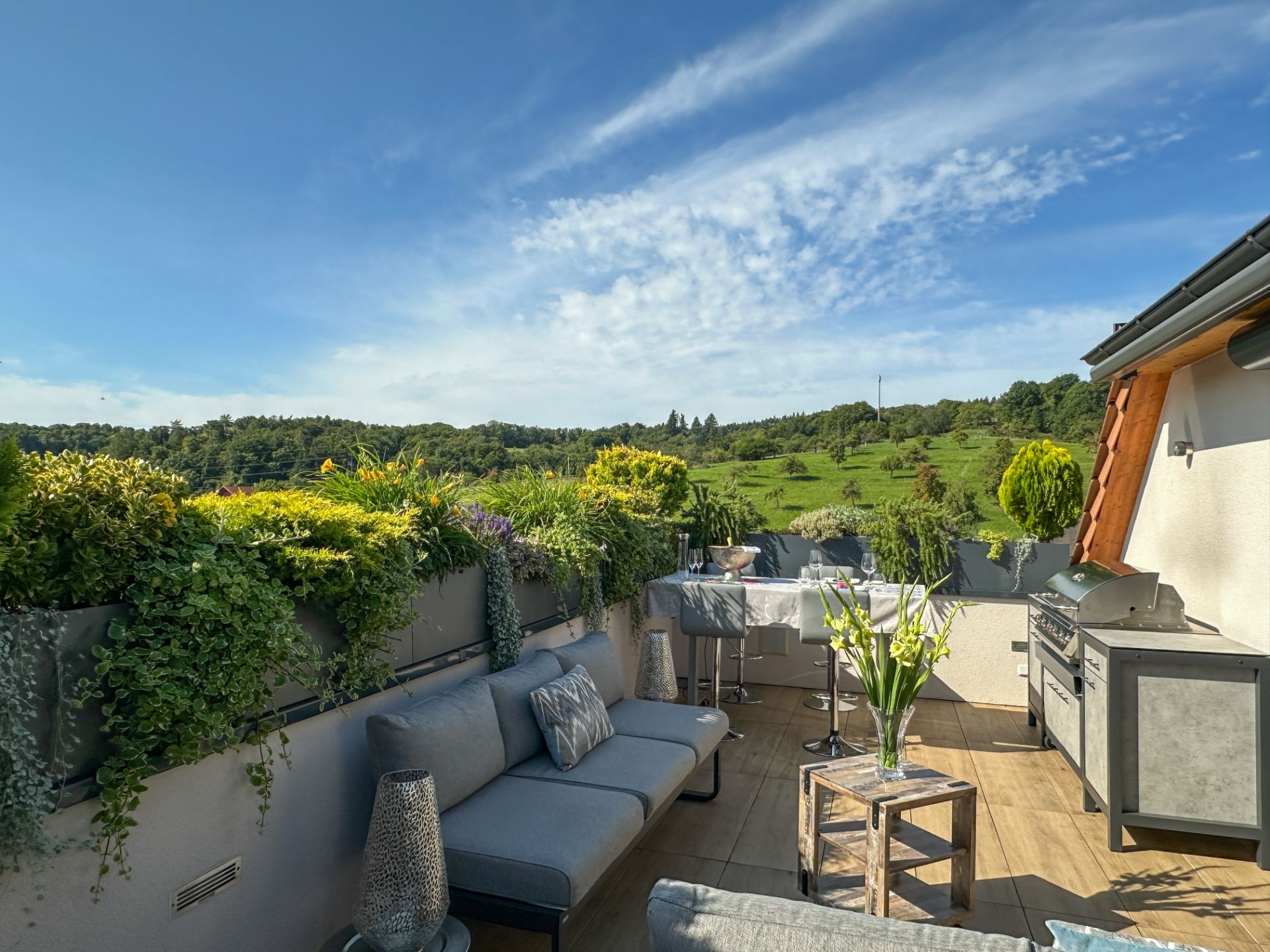 Moderne Dachterrasse mit Sitzmöbeln, Grill, Pflanzen und Ausblick ins Grüne.