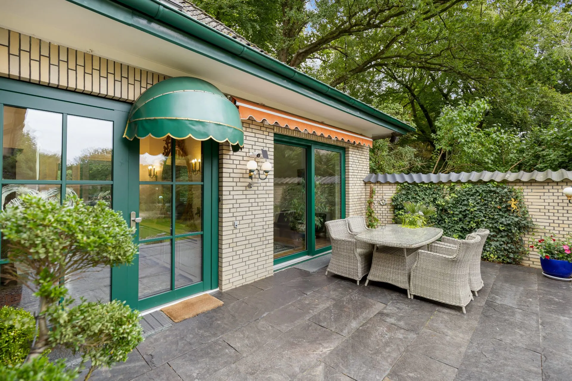 Terrasse mit Sitzgruppe, grüner Tür, Markise und bepflanzter Mauer im Hintergrund.