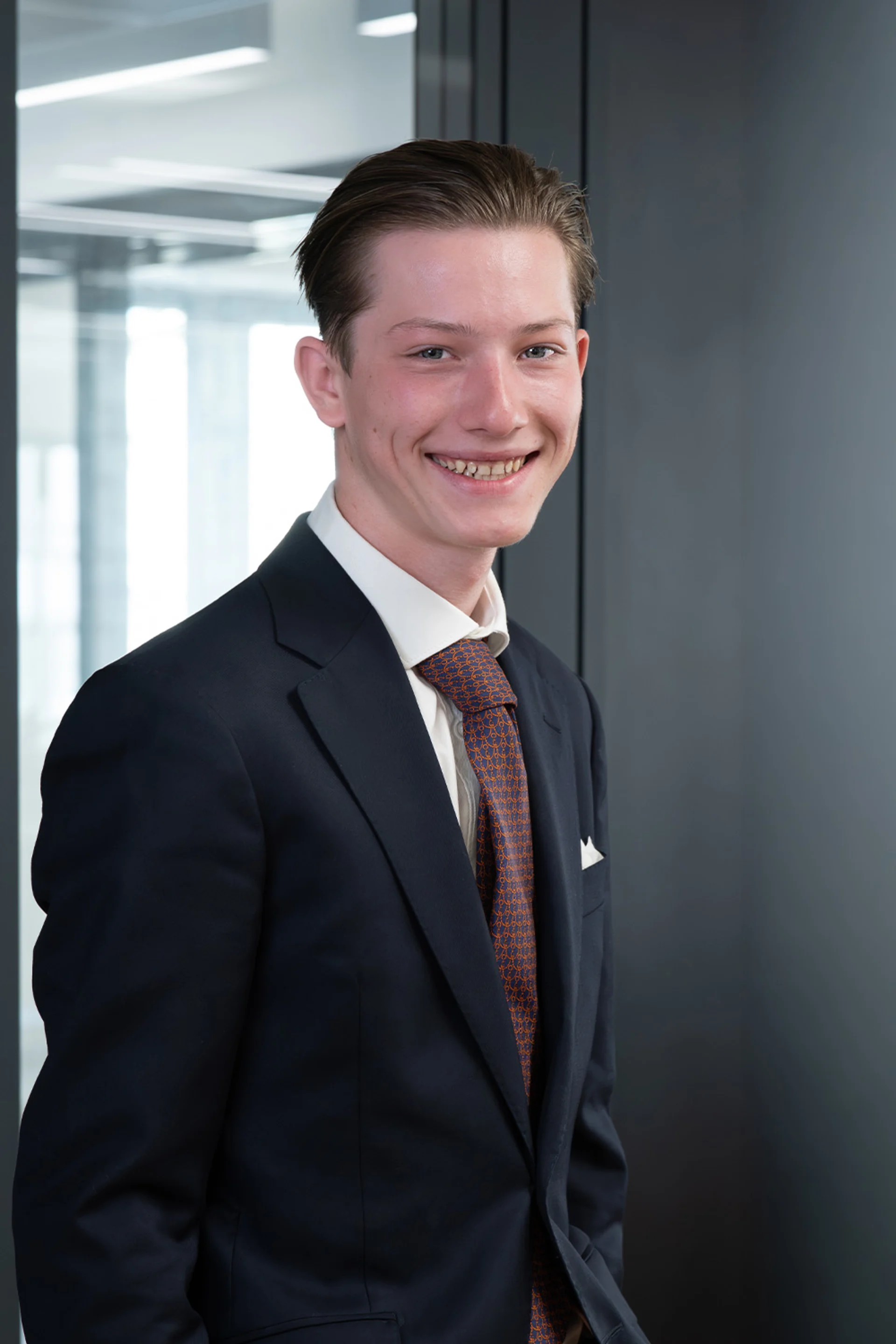 Junger Mann im Anzug mit Krawatte steht und lächelt in einem modernen, hellen Raum.