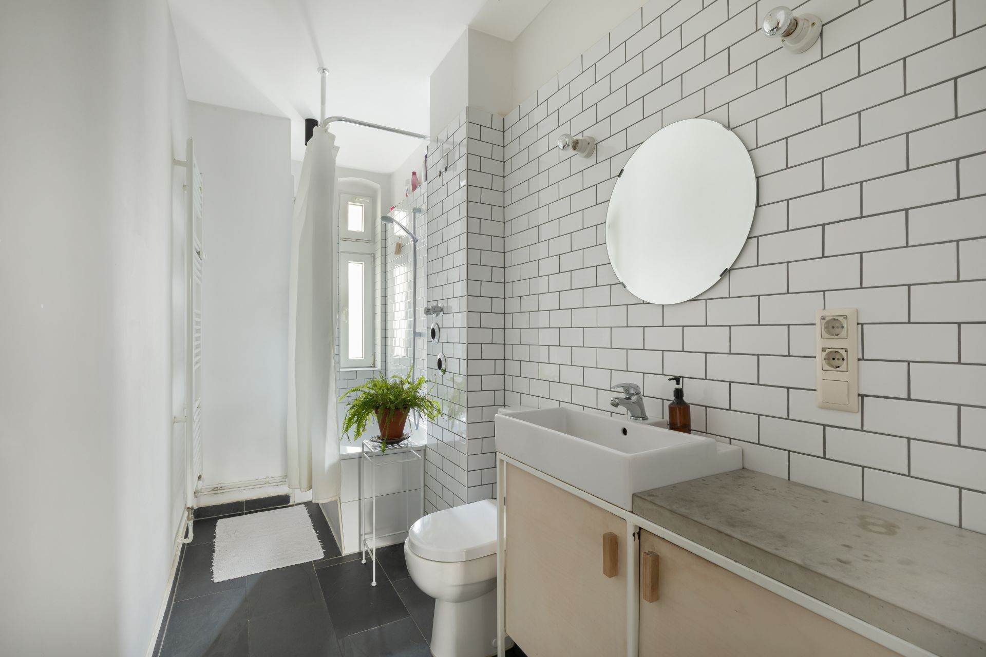 Helles Badezimmer mit weiß gefliester Wand, Waschbecken, rundem Spiegel und Dusche.