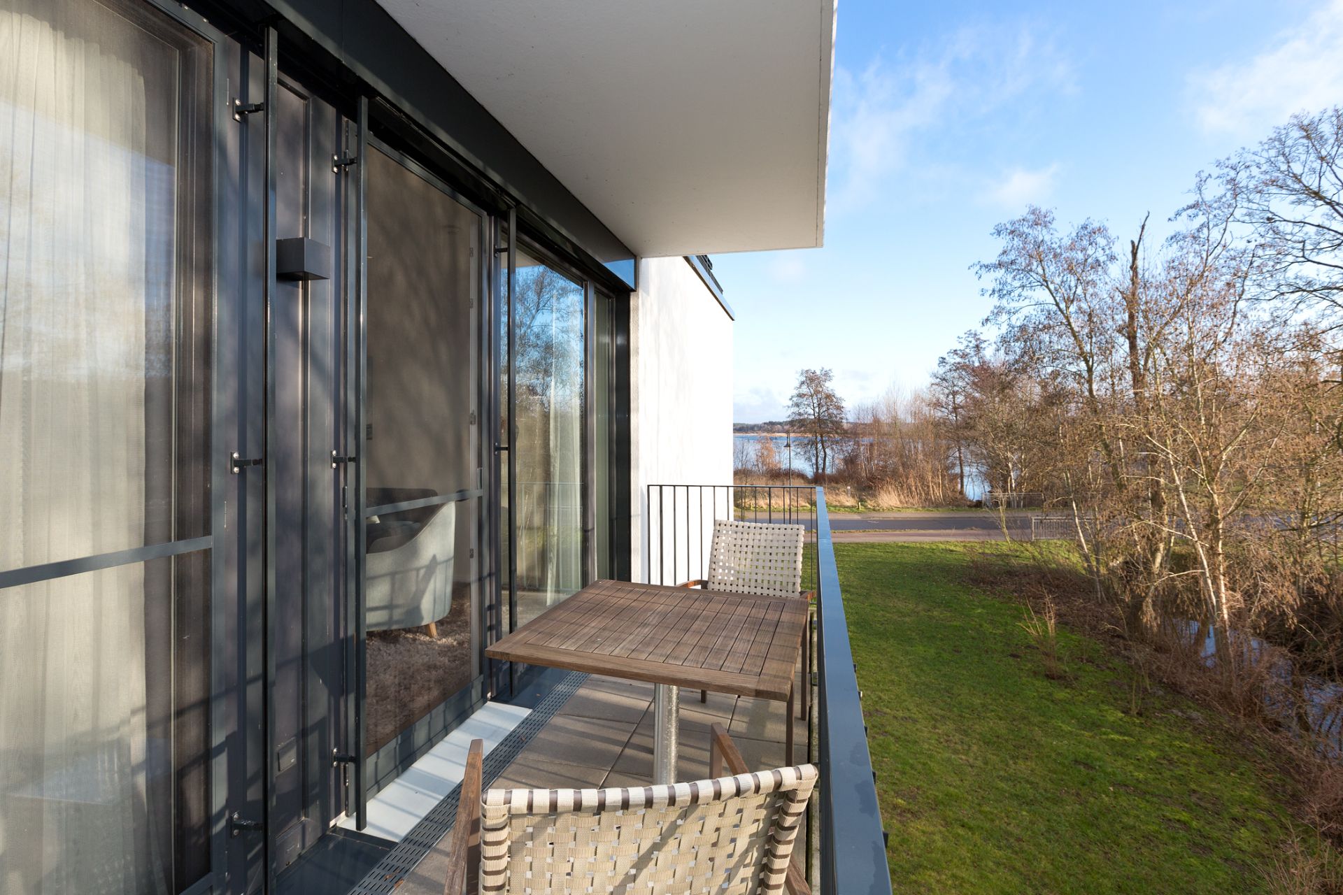 Balkon mit Holztisch und zwei Stühlen, Blick auf Wiese, Bäume und See im Hintergrund.