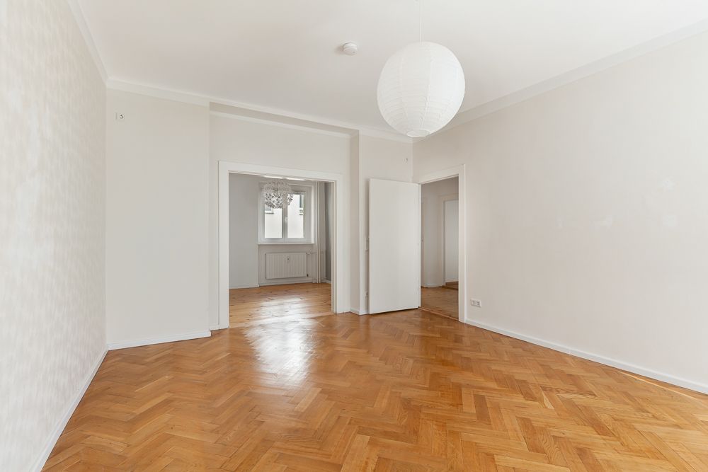 Leerer Wohnraum mit Parkettboden, weißen Wänden und offener Tür zu weiterem Zimmer.