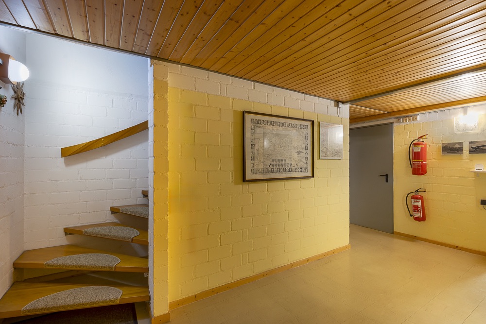 Flur mit Holzdecke, Treppe nach oben, gelbe Wände, Bildern und zwei roten Feuerlöschern an der Wand.