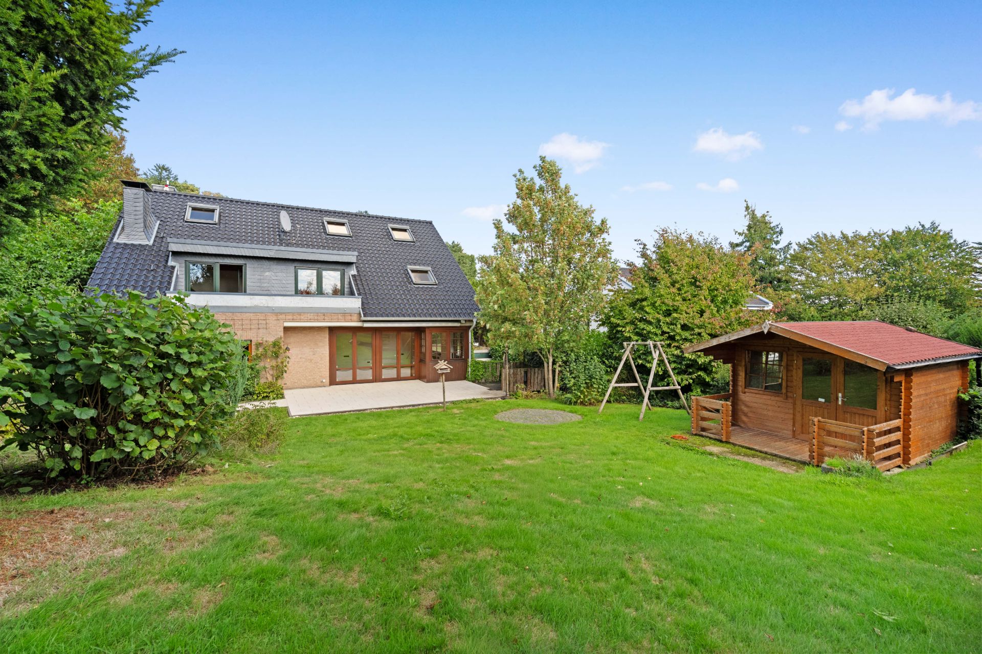 Haus mit Terrasse, großem Garten, Spielplatzschaukel und kleinem Holzhaus im Grünen.