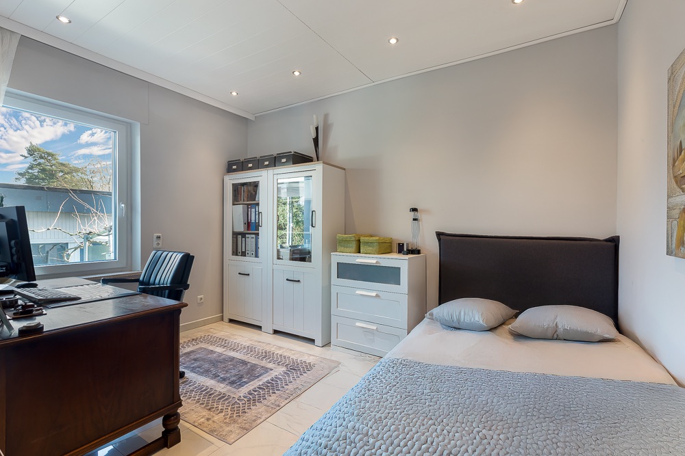 Helles Schlafzimmer mit Bett, Schreibtisch, Stuhl, Teppich und Schrank am Fenster.