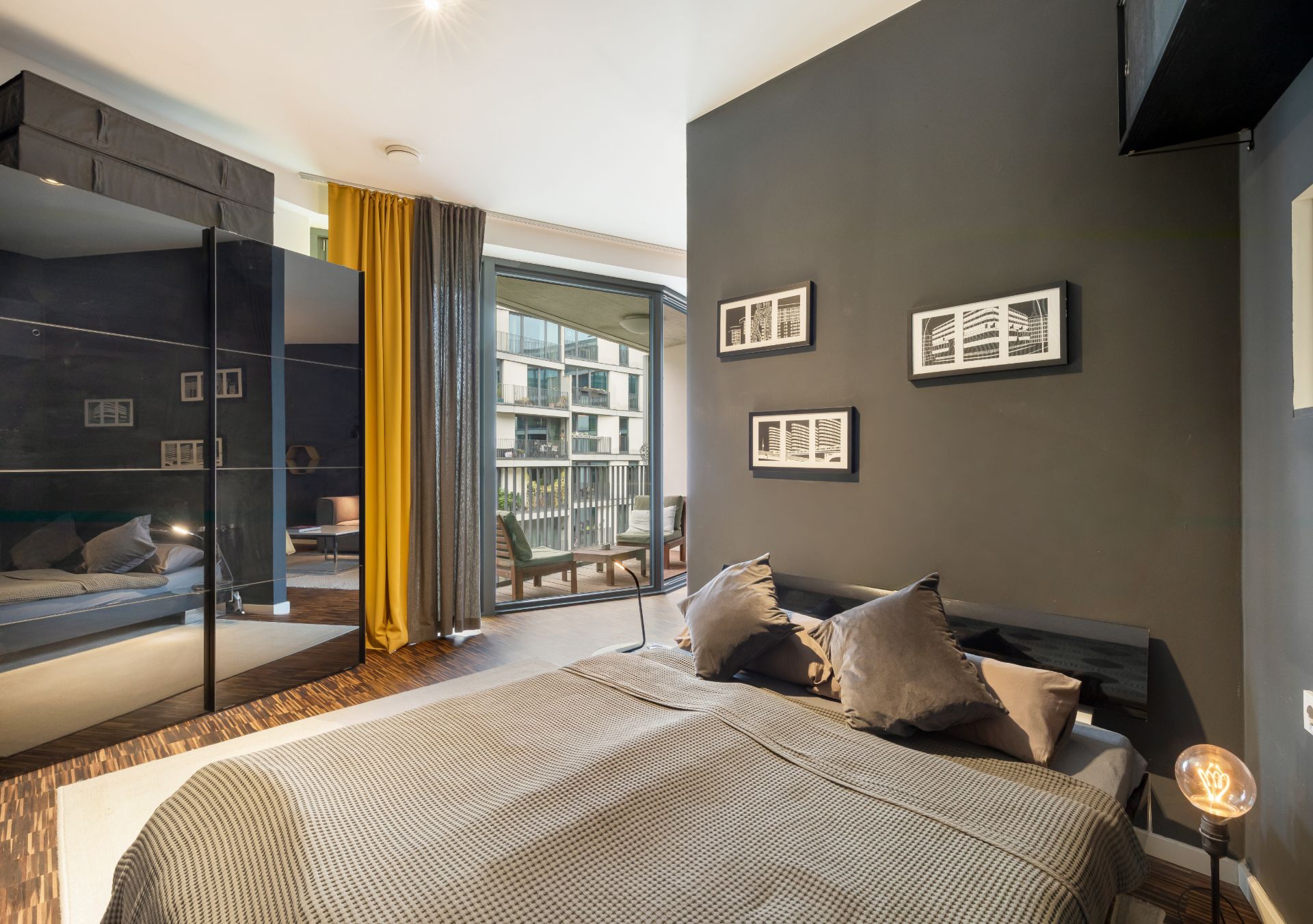 Modernes Schlafzimmer mit grauen Wänden, großem Bett, Spiegel-Kleiderschrank und Zugang zu einem Balkon.