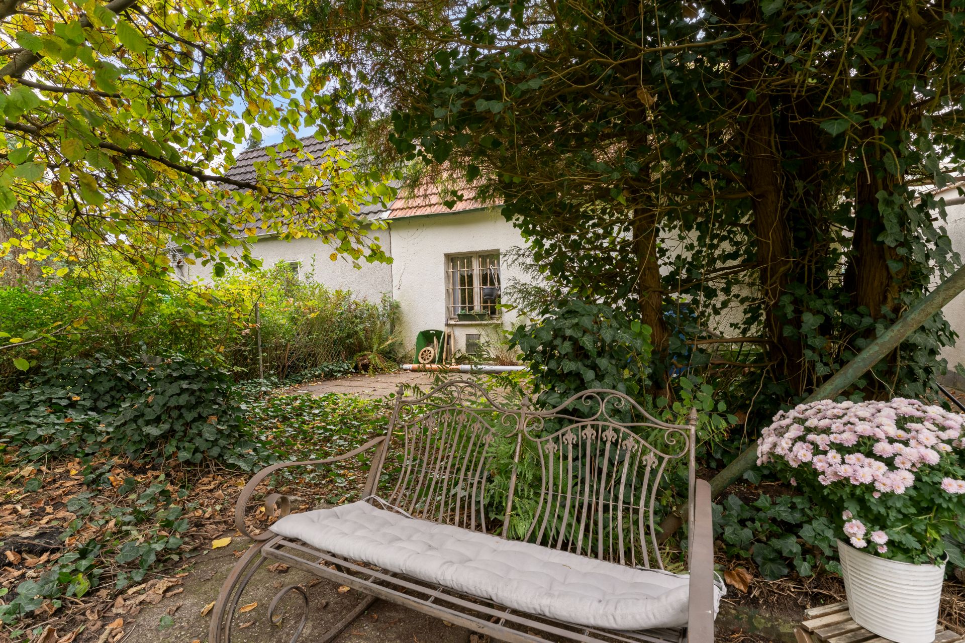 Metallbank mit Polster im Garten, große Büsche und Bäume, Haus im Hintergrund.