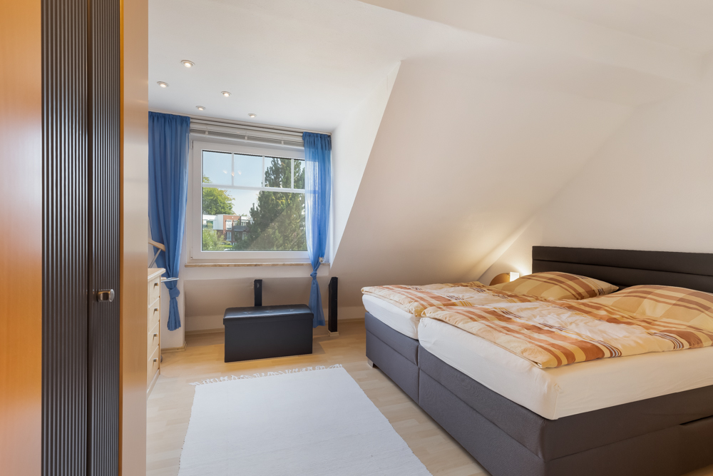 Helles Schlafzimmer mit Doppelbett, hellen Möbeln und großem Fenster mit blauen Vorhängen.
