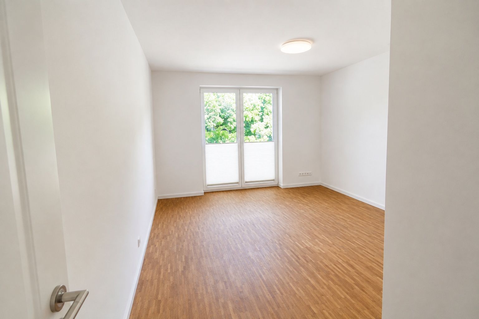 Helles, leeres Zimmer mit Holzfußboden und großen Fenstertüren zu einem Balkon.