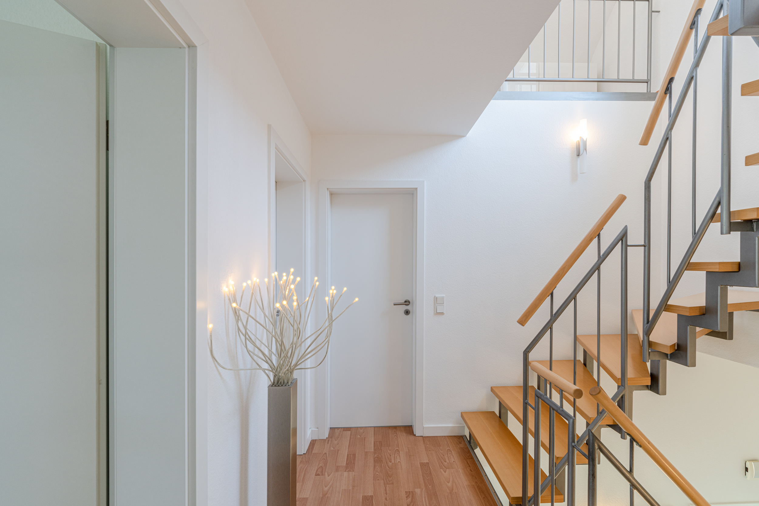 Heller Flur mit Holztreppe, weißer Tür und beleuchteter Dekoration in der Ecke.