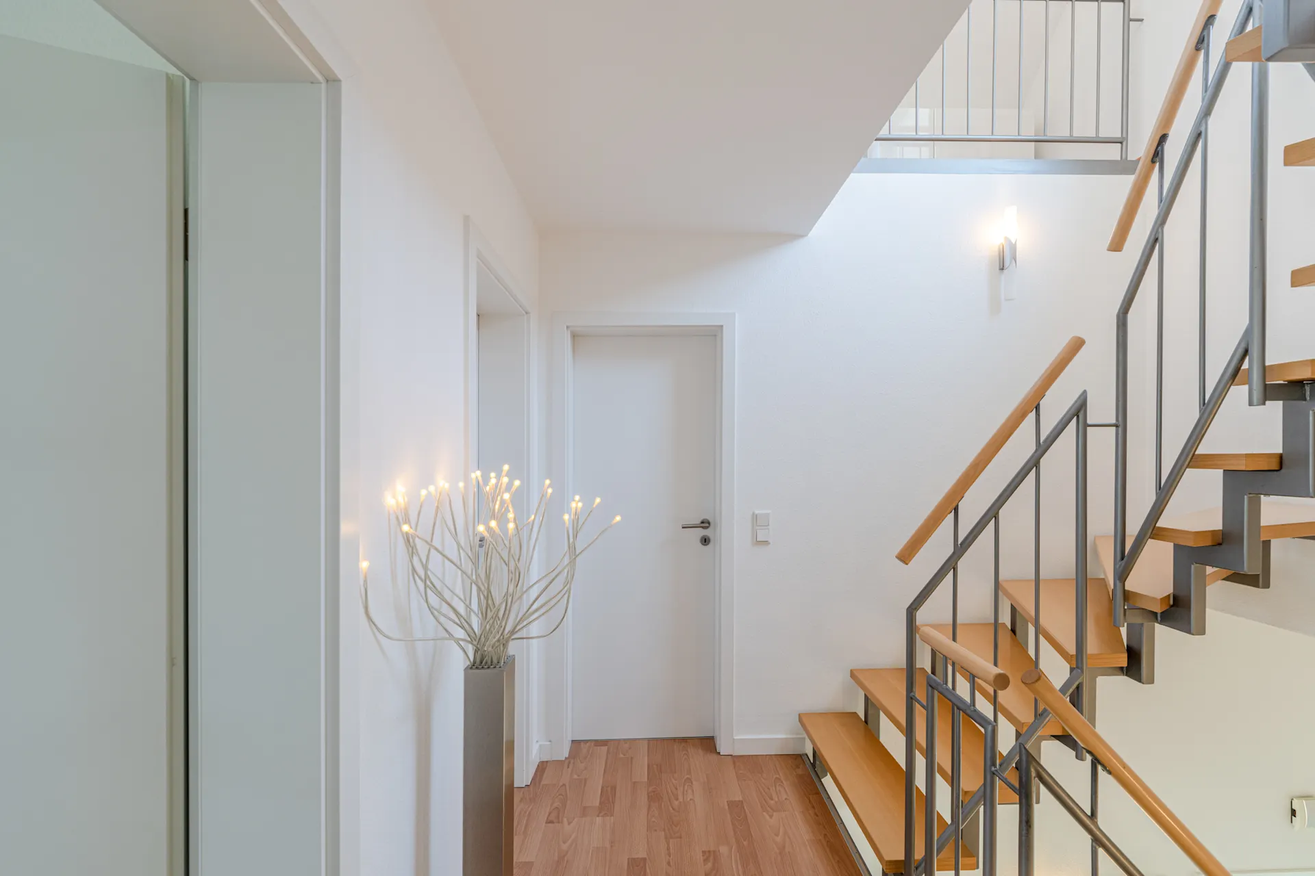 Heller Flur mit Holztreppe, weißer Tür und beleuchteter Dekoration in der Ecke.