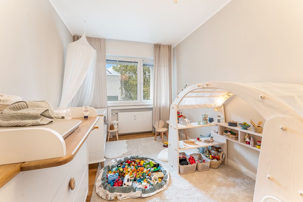 Helles Kinderzimmer mit Spielzeugteppich, Spielhaus, Wickelkommode und großem Fenster.