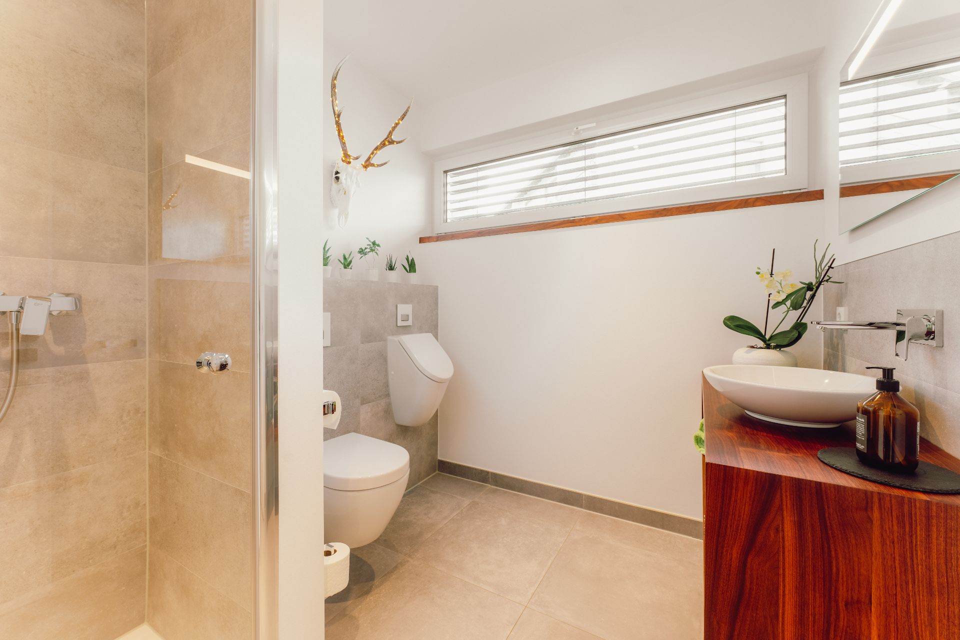 Modernes Badezimmer mit Dusche, WC, Urinal, Waschbecken und großem Fenster.